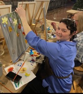 young female artist in a blue shirt seated at her painting easel with a painting of colorful flowers