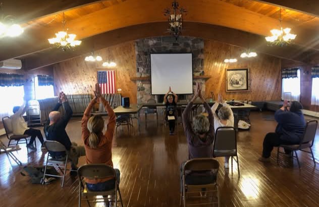 group of people seated in a large room following instructions by a person conducting a mindful movement exercise