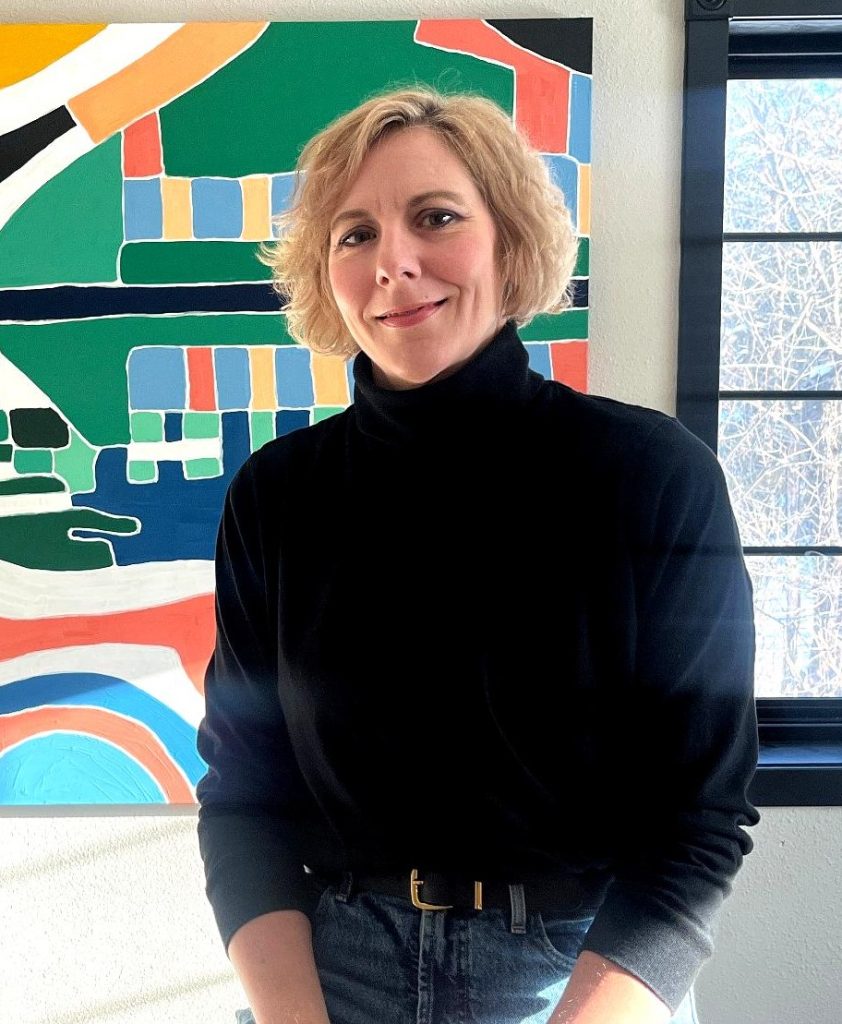 A smiling, blond haired adult female in a black turtleneck sweater standing in front of a colorful abstract painting.